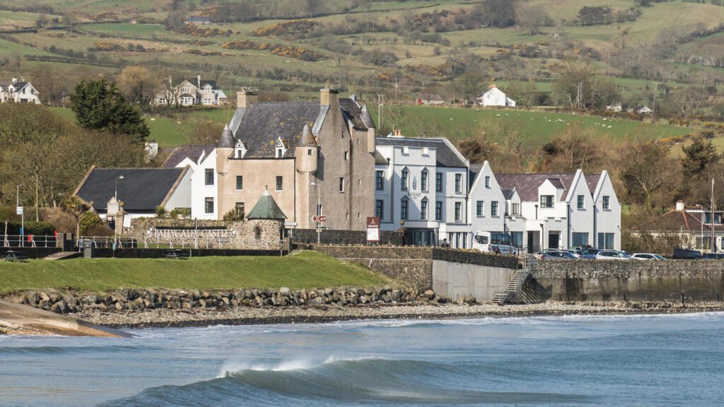 Ballygally Castle in Ireland, standing majestically behind a gently lapping body of water, with waves rolling in and a lush green hilly village in the background.