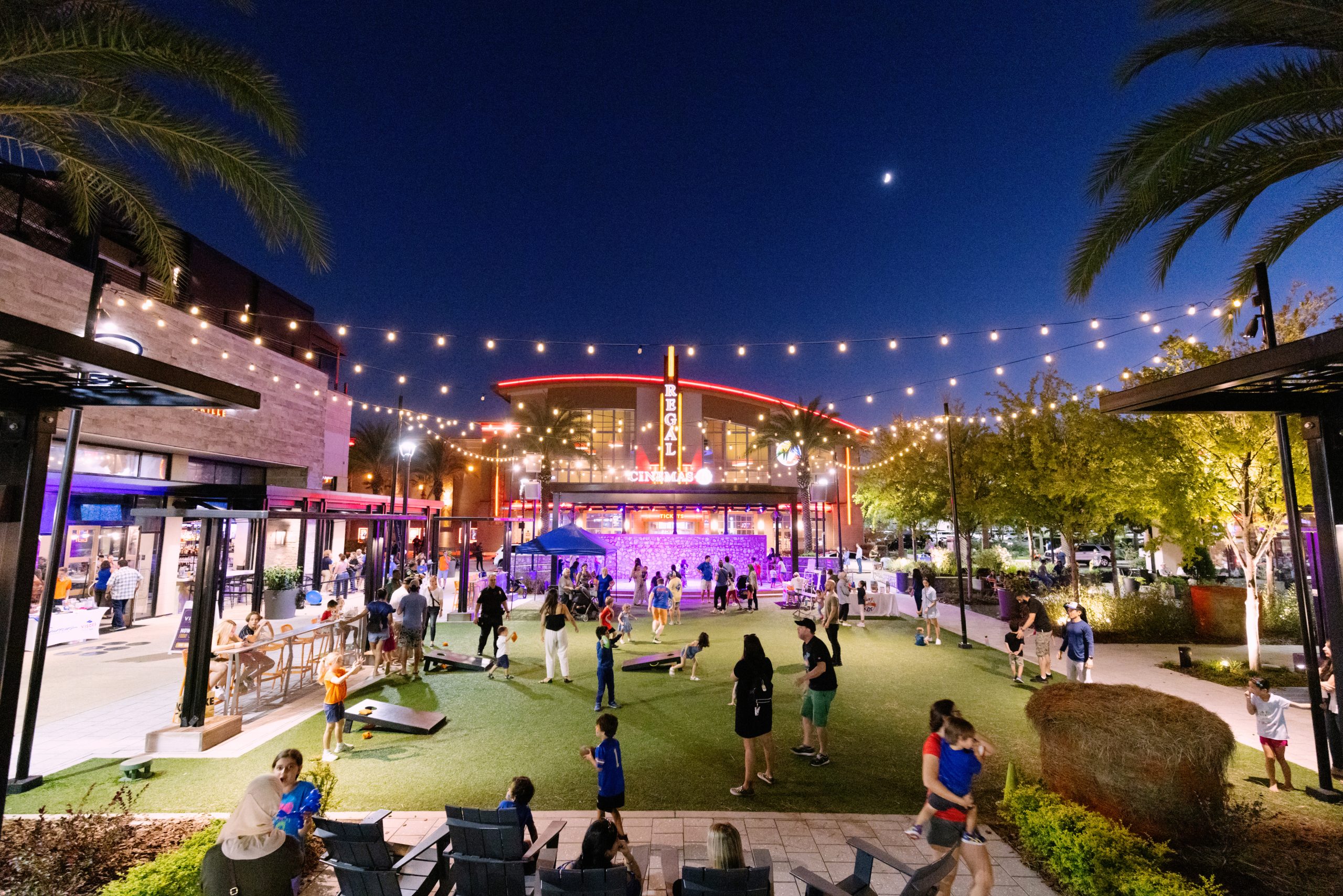 Nighttime view of Celebration Pointe in Gainesville, FL featuring an outdoor space lined with shops and a Regal Movies illuminated in the background, creating a vibrant atmosphere.