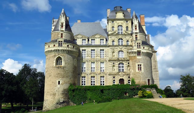 Château de Brissac, a stunning castle with a green lawn and ivy-covered front wall, set against a clear blue sky with fluffy white clouds.