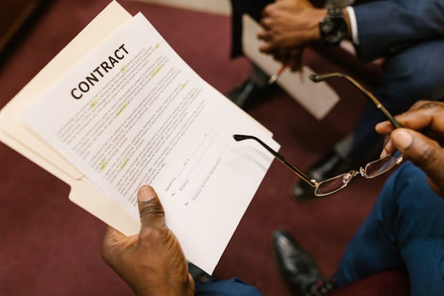 A man holding a contract and folder in his left hand while holding glasses in his right, with another man's hands blurred in the background, emphasizing a professional discussion about real estate.