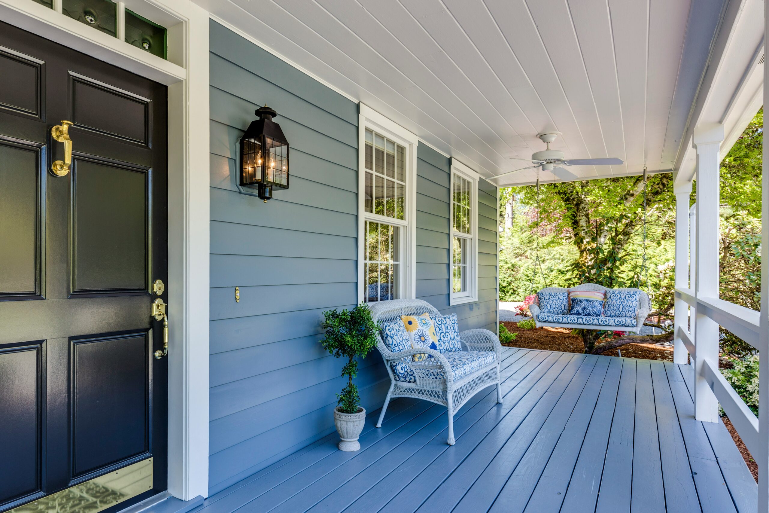 Charming porch featuring blue walls and floor, a white ceiling, with a cozy settee under two windows, and a porch swing in the background, complemented by a dark blue front door with gold accents.