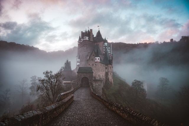 Fog-covered pathway leading to a large, historic castle with hints of blue sky above, setting a mysterious daytime atmosphere.