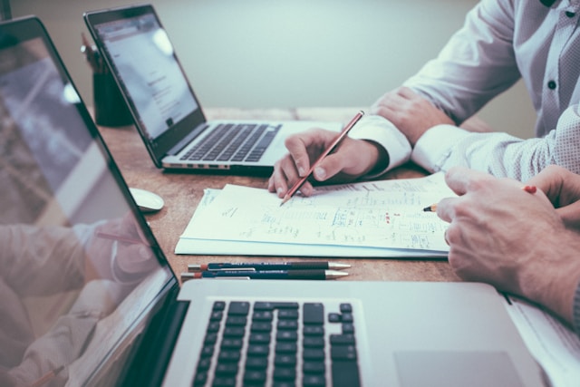 Two professionals at a table with two laptops and paperwork between them, both pointing to the documents, reflecting collaboration on real estate transactions or property management.