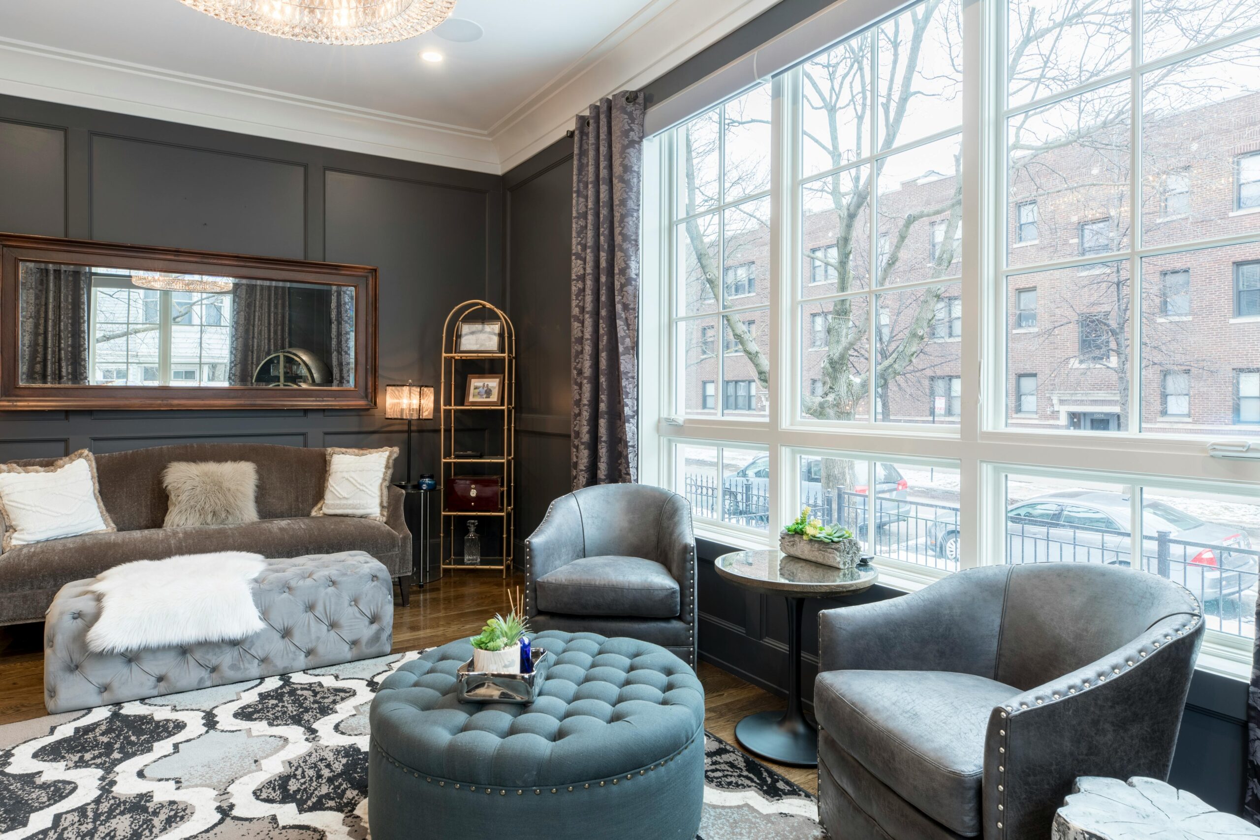 Classy living room with rich furnishings, including a couch and long ottoman beneath a decorative mirror, a chic rug, and a sitting area with two chairs and a round ottoman in front of a wall of windows.