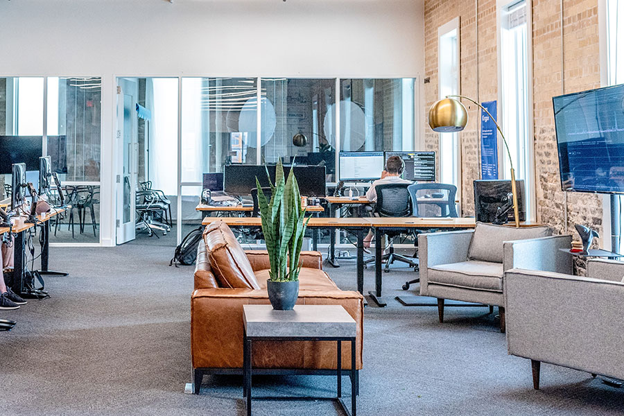 Modern open office featuring a sitting area with a light brown leather couch and two chairs in the foreground, with a row of desks and monitors in the background, including a person seated with their back facing us, and a glass wall of leading to an office.