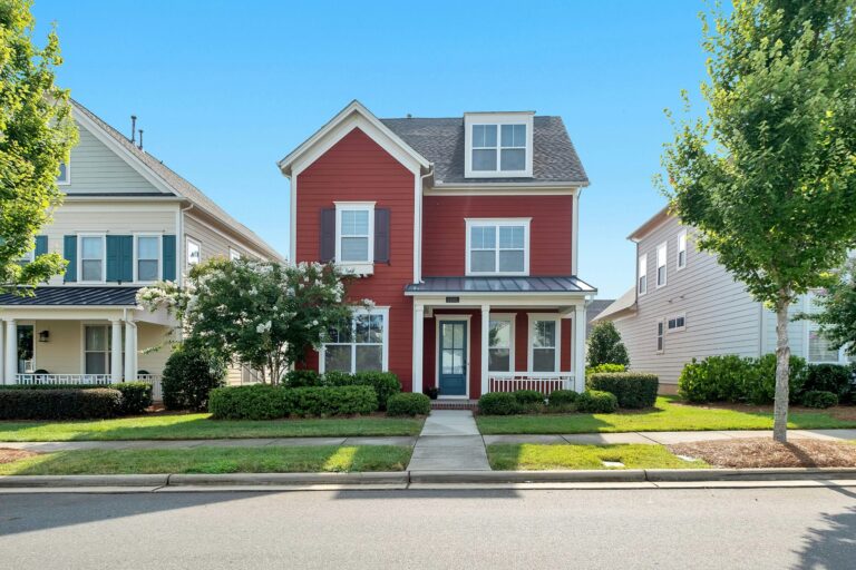 Street with three homes, a two-story red house is in the center featuring white trim, blue shutters and door, and landscaped yard, with partial views of neighboring houses.