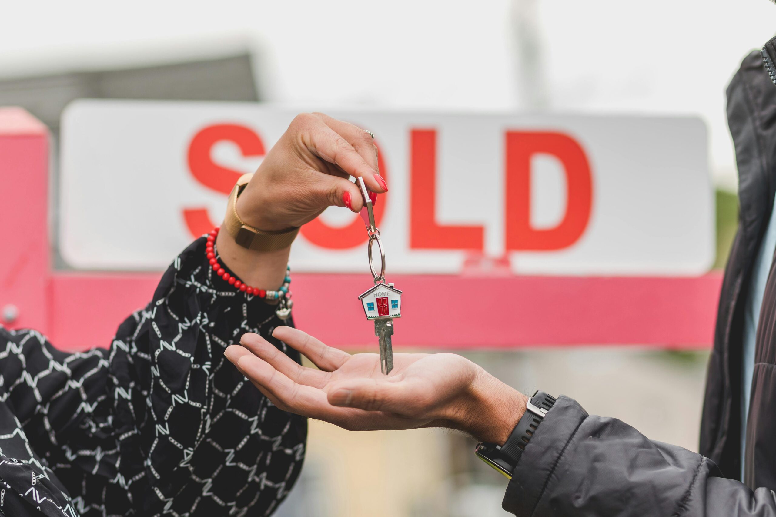 Close-up of a hand holding a key ring with a house keychain, another hand underneath, with a SOLD sign in the background, symbolizing a successful home sale.