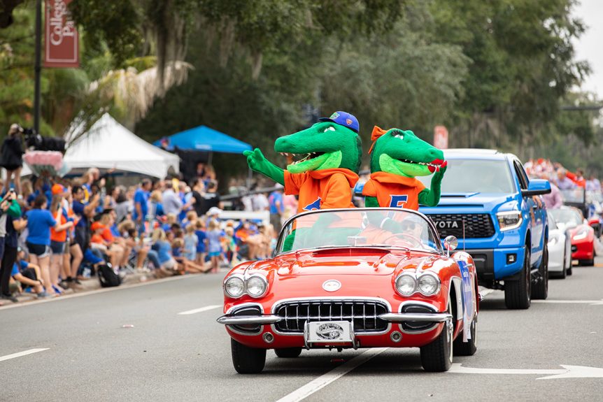 University of Florida mascots, Albert and Alberta, riding in a convertible during the UF homecoming parade, with a line of cars behind and spectators gathered on the left side.